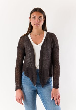 Giovane donna che indossa un cardigan marrone scuro trasparente aperto sul davanti sopra una maglietta bianca con scollo a V e jeans azzurri, in piedi davanti a uno sfondo neutro.