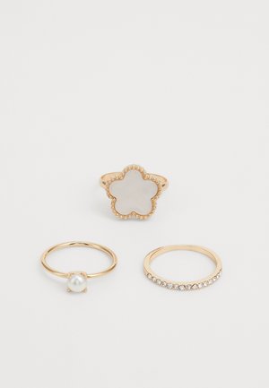 Trois bagues : une avec une base en or en forme de fleur et des accents en cristal, une avec une perle, et une avec une rangée de petites pierres transparentes.