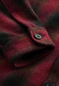 Poignet de chemise en flanelle à carreaux rouges et noirs avec un bouton noir, texture douce et ourlet roulé, idéal pour le superposition.