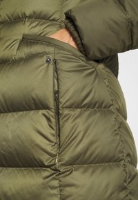 Giacca a vento verde oliva caratterizzata da sezioni trapuntate, una tasca con zip e un polsino lavorato a maglia visibile dalla manica.