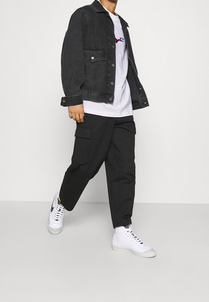 Man walking wearing a black denim jacket, white graphic t-shirt, black cargo pants, and white high-top sneakers.