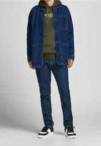 Giubbotto di jeans blu scuro con due tasche frontali, indossato sopra un hoodie verde con logo bianco, abbinato a jeans scuri e sneakers.
