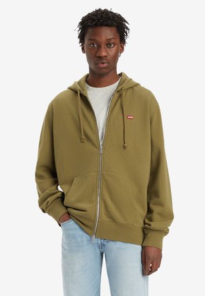 Sudadera con cremallera en color verde oliva, con un bolsillo tipo canguro, capucha ajustable con cordón y un pequeño logotipo rojo en el pecho, superpuesta sobre una camiseta gris.