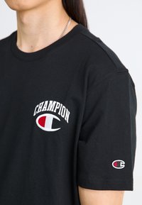 Melna kokvilnas T-krekls ar baltu izšūtu "CHAMPION" logotipu un sarkanu un baltu logotipu labajā piedurknē. Īsas piedurknes un apļa kakla izgriezums.