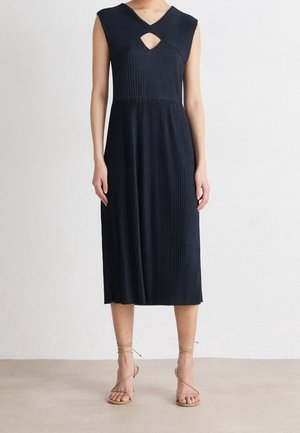 Femme portant une robe midi noire sans manches en maille côtelée avec un col en V et une petite découpe à l'avant, associée à des sandales beige à brides.