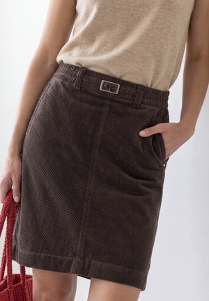 Mujer con una blusa beige y falda de pana marrón oscuro con cinturón, sosteniendo un bolso de punto rojo, mano en el bolsillo de la falda.