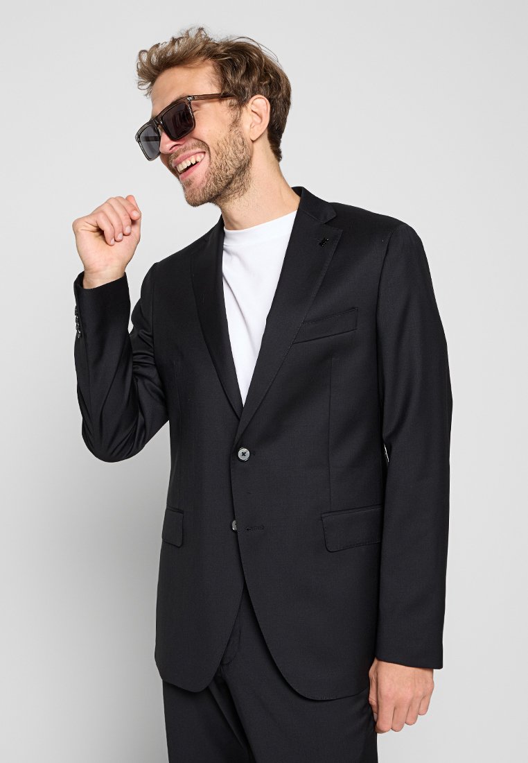 Man in een zwart pak, wit shirt en grote donkere zonnebril, lachend en met een hand bij zijn gezicht tegen een effen achtergrond.