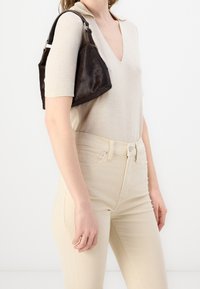 Sac à main en tissu synthétique marron tenu par une personne, associé à un haut beige à col en V et un jean ajusté beige clair.