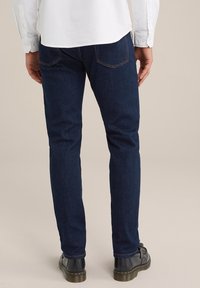 Sötétkék, slim fit farmernadrág, két hátsó zsebbel és kontrasztos varrással, fekete bőr cipővel párosítva.
