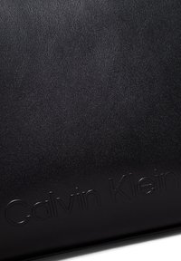 Czarny skórzany dodatek o teksturowanej powierzchni i wytłoczonym logo "Calvin Klein". Elegancki design z minimalistycznymi akcentami.