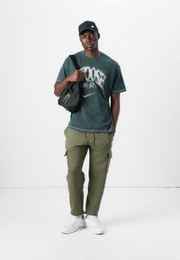T-shirt tinto di verde con testo grafico, pantaloni cargo oliva, borsa a tracolla nera e scarpe sportive bianche. Un cappellino nero completa l'outfit.