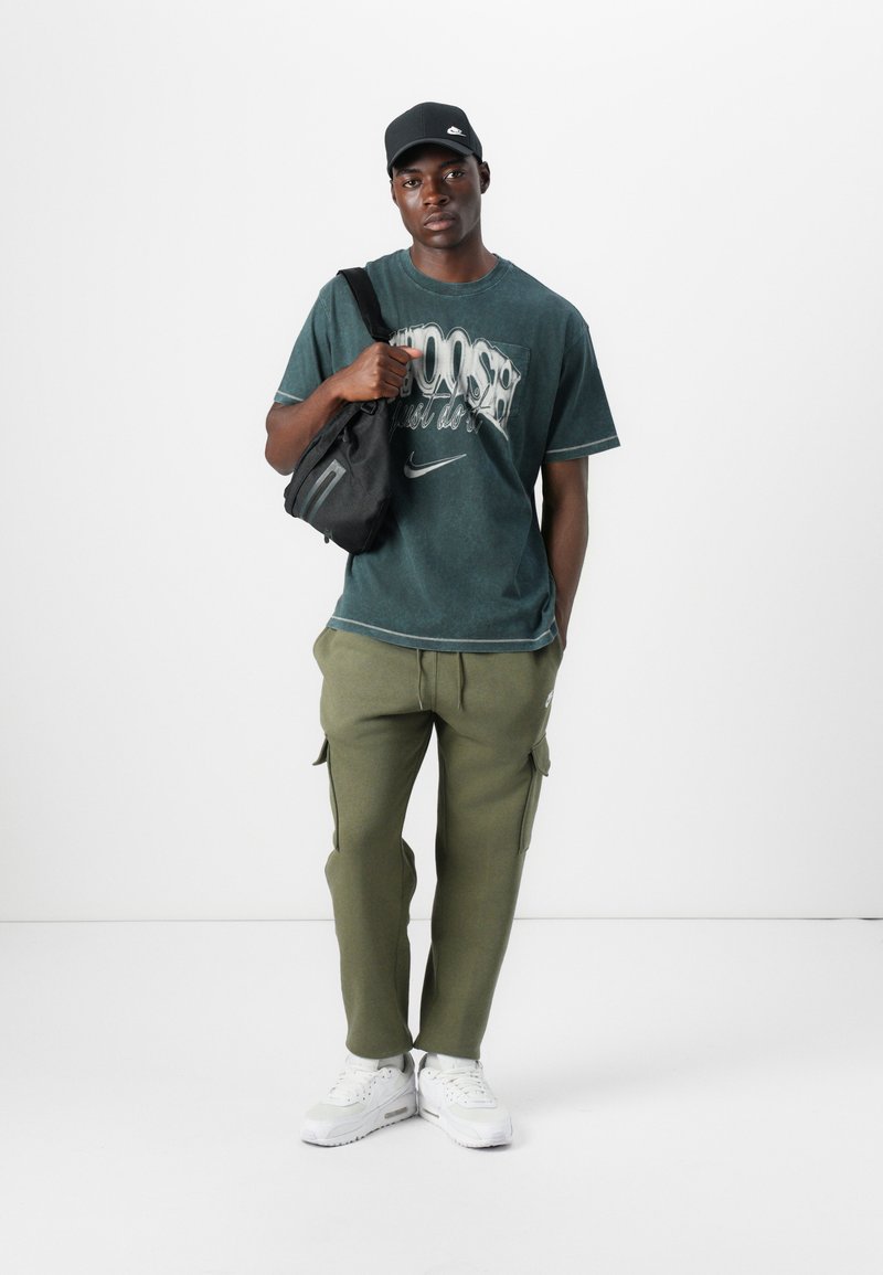 Grün gefärbtes T-Shirt mit grafischem Text, olivfarbene Cargo-Hosen, schwarze Umhängetasche und weiße Sportschuhe. Eine schwarze Cap rundet den Look ab.