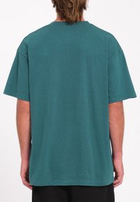 Un adulte portant un t-shirt simple teal surdimensionné à manches courtes, vu de dos avec les mains détendues sur les côtés.