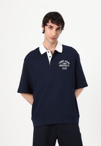Sötétkék póló rövid ujjakkal, fehér gallérral, az elején fehéren a "TOMMY JEANS UNIVERSITY CLUB" logóval.