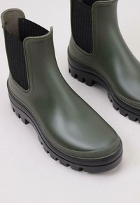 Botas de tobillo de goma verde con panel lateral de tela negra texturizada, suela gruesa con relieve y acabado suave. Apta para condiciones húmedas.