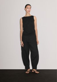Een vrouw staat zelfverzekerd tegen een lichte achtergrond, gekleed in een zwart mouwloos shirt en losse zwarte broek, met blote voeten en minimale accessoires.
