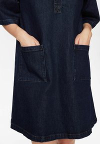 Donkerblauwe denim jurk met een losse pasvorm, voorzien van twee voorzakken en een lichte curve aan de zoom. Knopen bij de halslijn.