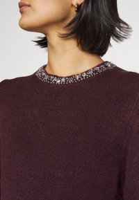 Femme portant un pull en tricot violet foncé avec un col orné de perles et de bijoux et de petites boucles d'oreilles en or, vue du cou aux épaules.