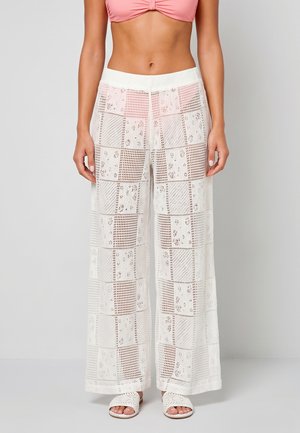 Vrouw draagt witte doorschijnende gehaakte broek met wijde pijpen, roze cropped top en witte geweven slippers, staand tegen een effen achtergrond.