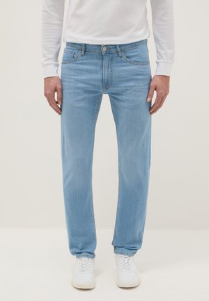 Mann, der hellblaue Jeans mit geradem Bein, ein weißes Langarmhemd und weiße Turnschuhe trägt und vor einem einfarbigen Hintergrund steht.