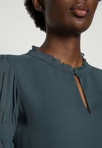 Blouse bleu-vert avec un col froncé, un décolleté en forme de clef, et des manches bouffantes. Le tissu a une finition texturée avec des lignes verticales subtiles.