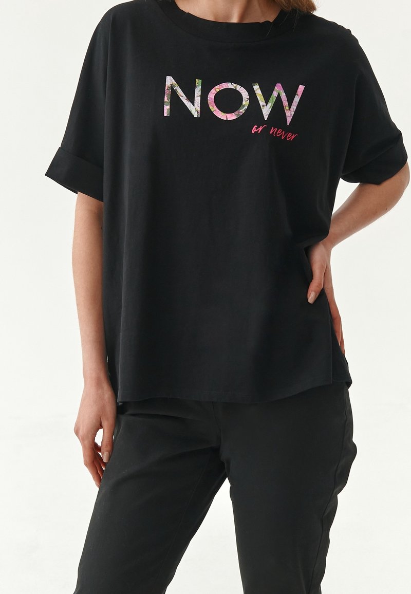 Personne portant un t-shirt noir ample avec le texte « NOW or never », associé à un pantalon noir, main posée sur la hanche, fond uni.