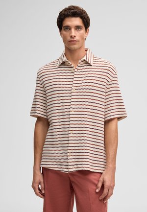 Mężczyzna ubrany w koszulę na krótki rękaw zapinaną na guziki z poziomymi, czerwono-białymi falistymi paskami oraz rdzawobrązowe spodnie, stojący na jednolitym tle.