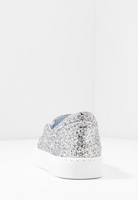 Silver glittrande sneakers med vit sula, visad från bakvinkel på en vit yta mot en slät vit bakgrund.