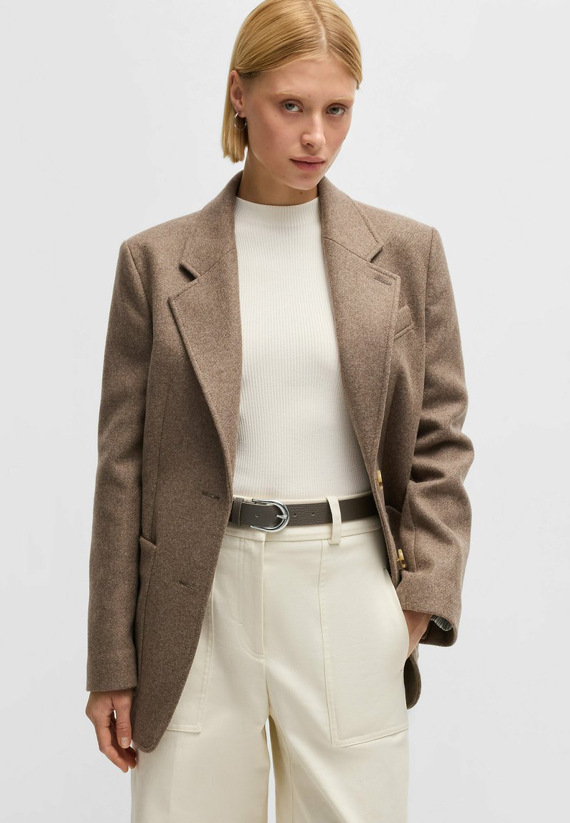 Brun kashmirblandad kavaj med notsch krage, dubbelknäppt design och knappar i guldton, kombinerad med en vit ribbad topp och beige byxor.