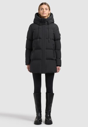 Femme portant un manteau matelassé noir à capuche avec poches zippées, des leggings noirs et des bottes noires montantes jusqu'aux genoux, debout devant un fond clair uni.