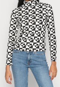 Långärmad polokrage topp med ett svartvitt hjärtmönster på en rutigt bakgrund, tillverkad av stretchigt tyg. Kombinerad med blå jeans.