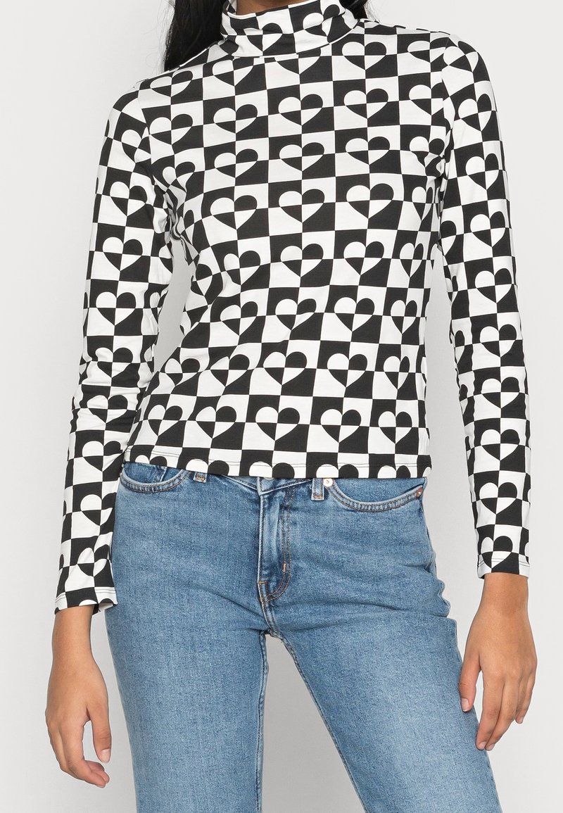 Långärmad polokrage topp med ett svartvitt hjärtmönster på en rutigt bakgrund, tillverkad av stretchigt tyg. Kombinerad med blå jeans.