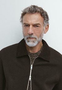 Giacca marrone conzip e colletto, indossata sopra un maglione marrone a trama. Capelli corti e ricci e barba grigia. Sfondo neutro.