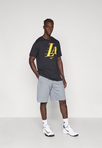 Musta T-paita, jossa keltainen "LA"-logo, harmaat shortsit ja valkoiset tennarit. Sileää kangasta, lyhyet hihat, rento istuvuus ja hillitty brändäys sukissa.