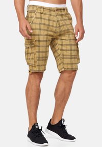 Cargo shorts in a tan color with a black plaid pattern. Features include cargo pockets, a button closure, and a relaxed fit design.