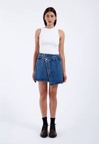 Denim-Minirock mit asymmetrischem Design, hellblauer Waschung, rohem Saum und einem einzelnen Knopfverschluss. Kombiniert mit einem weißen Tanktop und dunklen Schuhen.