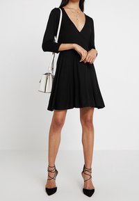 Svart knälång klänning med djup V-ringning och trekvartslånga ärmar. Tillverkad av mjukt tyg och har en utsvängd silhuett. Matchad med svarta remsandaler och en liten vit handväska.