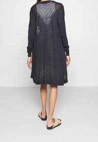 Lång mörk tröjdress med långa ärmar, öppen stickning på baksidan och en avslappnad passform, parat med ljusa slip-in sandaler.