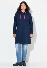 Abito felpa navy con zip frontale, cordini rosa, maniche lunghe e taglio dritto; abbinato a jeans azzurri chiari e stivali scuri.