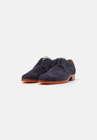 Scarpe in suede blu navy con una texture liscia, punta rotonda, lacci sottili e una suola in gomma arancione a contrasto. Presentano un piccolo accentuamento sul tallone.