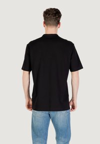 Camiseta negra de manga corta hecha de algodón, con cuello redondo y corte relajado, usada con jeans de color azul claro. Costuras visibles y etiqueta en el dobladillo.