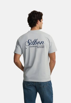 Hombre con cabello rizado y oscuro, lleva una camiseta Silbon gris clara y vaqueros azules, de espaldas mostrando el texto en la parte posterior de la camiseta.