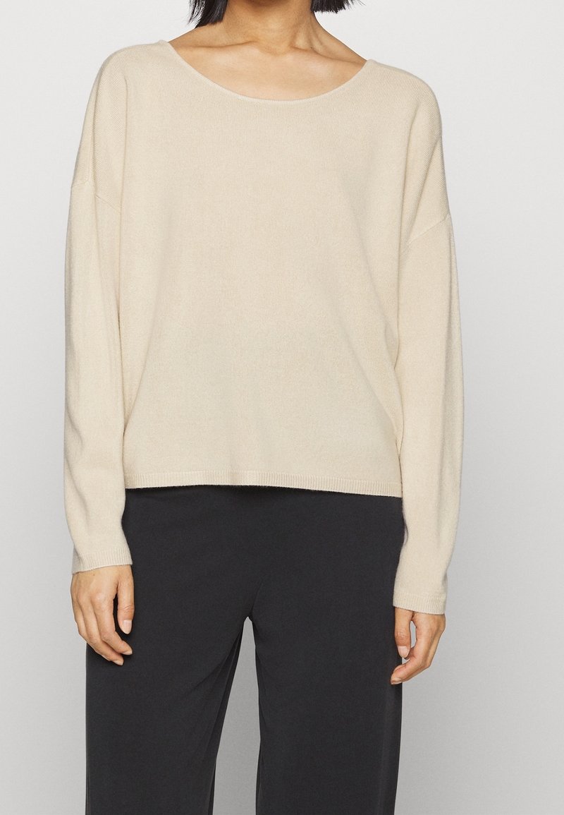 Beige sweater met ronde hals, lange mouwen, een zachte gebreide textuur en een ontspannen pasvorm, gecombineerd met zwarte broek voor contrast.