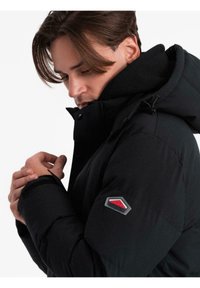 Uomo con giacca invernale nera con cappuccio che aggiusta la manica, mostrando una toppa logo rossa e grigia sul braccio superiore contro uno sfondo bianco.