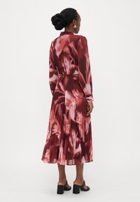 Mittelkurzes, plissiertes Kleid in Burgunderrot mit abstrakten pinken Mustern, langen Ärmeln und einem gebundenen Taillengürtel. Kombiniert mit schwarzen, hohen Sandalen.