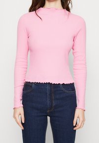 Långärmad topp i ljusrosa ribbat tyg, med en vågformad halsringning och nederkant, i kombination med mörkblå jeans.