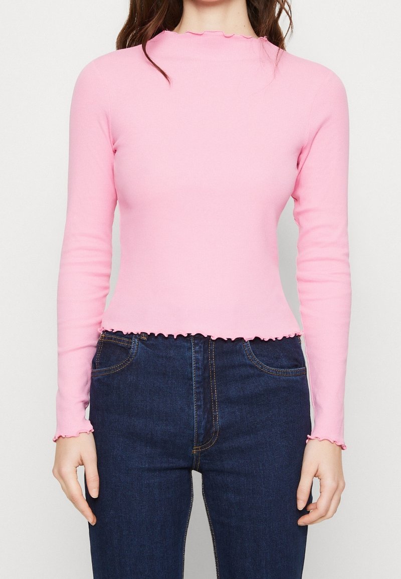 Långärmad topp i ljusrosa ribbat tyg, med en vågformad halsringning och nederkant, i kombination med mörkblå jeans.