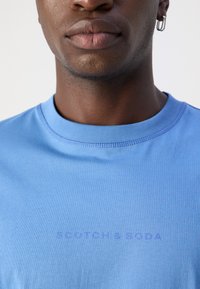 Gaiši zils kokvilnas T-krekliņš ar apaļu kakla izgriezumu, ar "SCOTCH & SODA" tekstu drukātā nedaudz tumšākā zilā krāsā, īsām piedurknēm un gludu tekstūru.