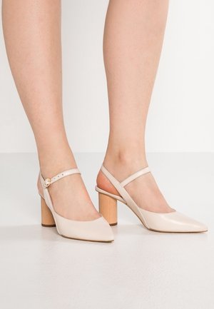 Escarpins slingback beige à bout pointu avec talons blocs en bois, portés pieds nus sur un fond clair.