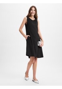 Zwarte mouwloze jurk van lichtgewicht stof, met zijzakken, een losse pasvorm en een ronde halslijn. Zilveren clutch en schoenen.